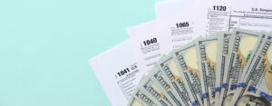 Tax forms lies near hundred dollar bills and blue pen on a light blue background.