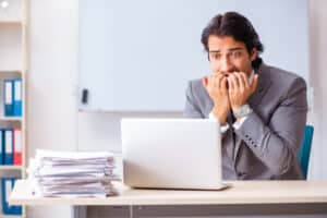 Grey and white image of a businessman looking at computer while holding his hands in front of his scared face. A pile of paperwork is on the side of the computer. 