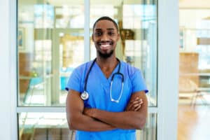 Image is a smiling male nurse practitioner wearing blue scrubs and a stethoscope.