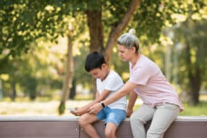 Mother trying to take a mobile phone away from her 10 years old son, sitting on the bench outdoor in public park in summer, selective focus. Digital addiction, games, parenting, child issues concept